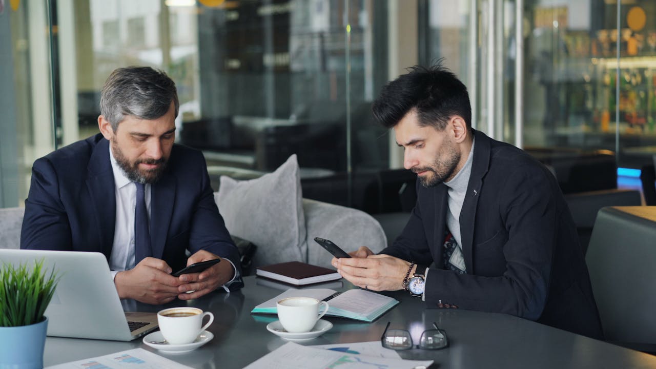 Two businessmen in a cafe working on smartphones and laptops, engaged in a meeting.